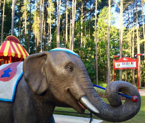 A large elephant statue with a blue hat, set in a wooded area near a colorful amusement park entrance.