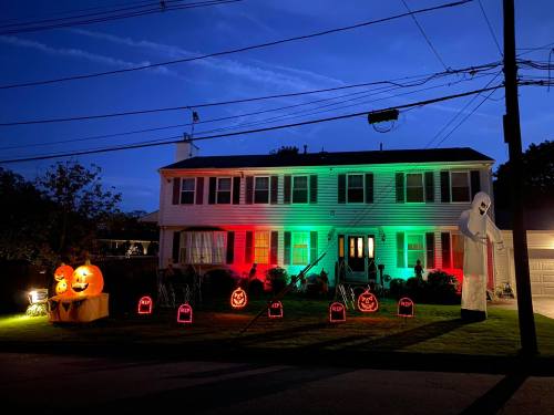 A house decorated for Halloween with green and red lights, pumpkins, tombstones, and a ghost figure in the yard.
