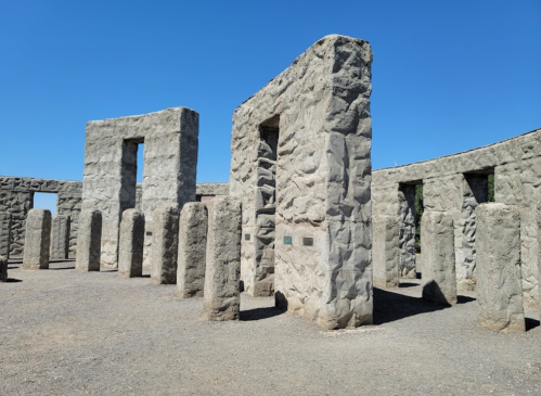 Stone structures arranged in a circular formation under a clear blue sky, with tall pillars and a gravel ground.