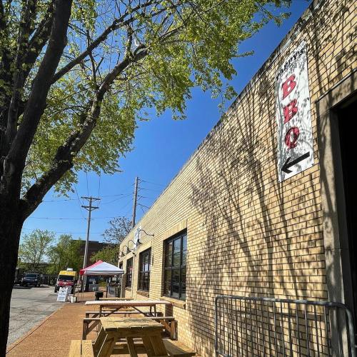 A sunny outdoor area with picnic tables, a tree, and a building featuring a BBQ sign.