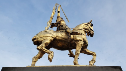 A golden statue of a mounted warrior holding a sword, set against a blue sky.