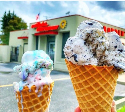Two ice cream cones in front of an ice cream shop, one with colorful sprinkles and the other with cookies and cream.