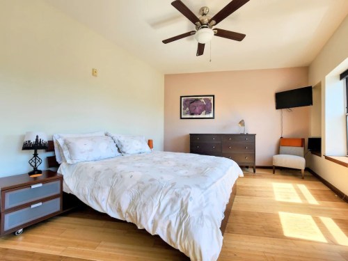 Bright bedroom with a queen bed, wooden furniture, a ceiling fan, and a TV mounted on the wall. Natural light floods the space.