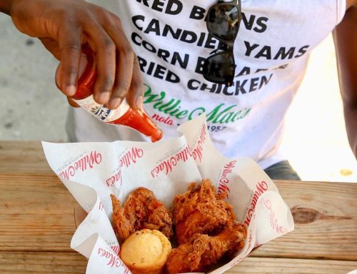 A person in a t-shirt pours hot sauce over fried chicken and cornbread on a wooden table.