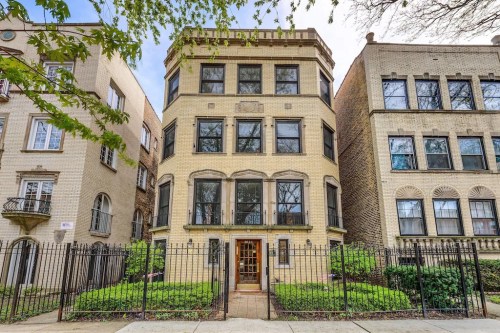 A three-story brick building with large windows, surrounded by greenery and a wrought-iron fence.
