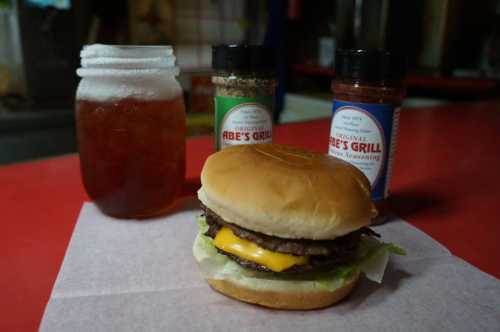 A cheeseburger with lettuce on a bun, accompanied by a jar of iced tea and seasoning bottles on a red table.