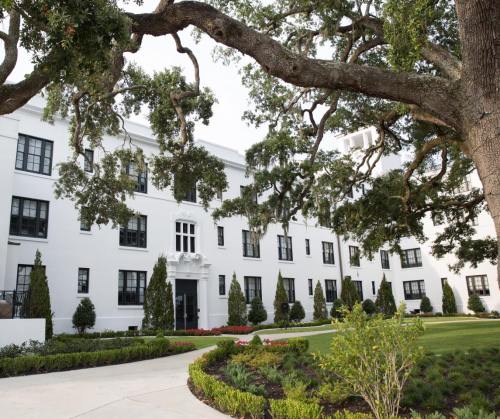 A white building with black windows surrounded by greenery and large trees, featuring a manicured lawn in front.