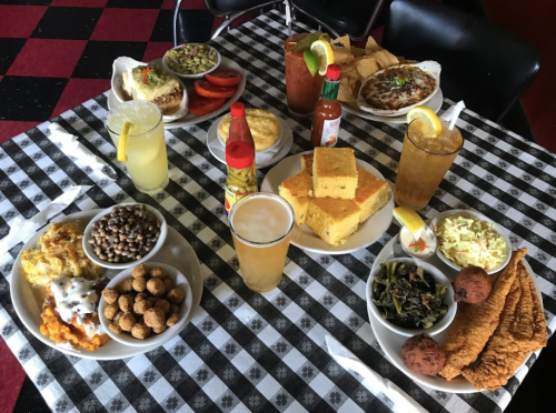 A spread of Southern dishes on a checkered tablecloth, featuring fried fish, cornbread, collard greens, and drinks.