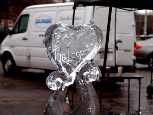 Ice sculpture in the shape of a heart with "I ❤️ The Loop" carved into it, set against a backdrop of vehicles.