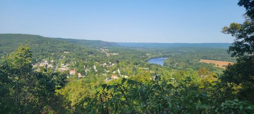 A panoramic view of a lush green valley with a river, small town, and distant hills under a clear blue sky.