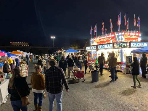 A lively night market scene with people walking around food stalls and colorful tents under a dark sky.