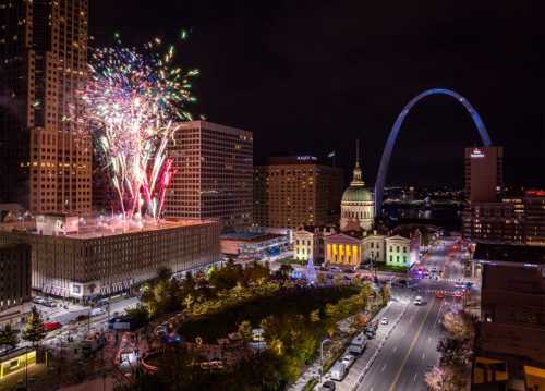 Colorful fireworks illuminate the night sky over a cityscape featuring an arch and festive buildings.