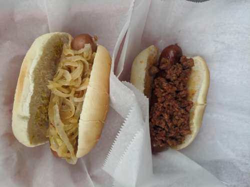 Two hot dogs on a paper wrapper: one topped with sauerkraut and mustard, the other with chili.
