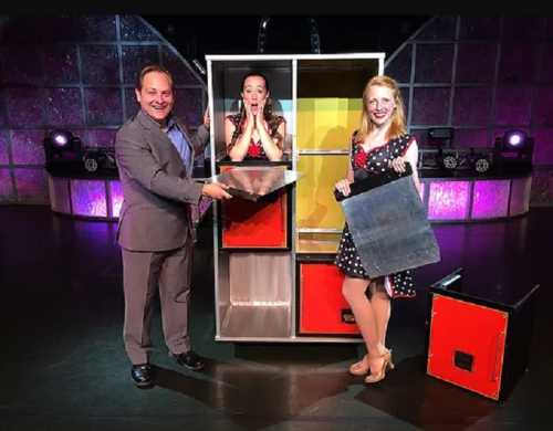 A magician and two assistants pose with a colorful magic box on stage, showcasing a playful performance.