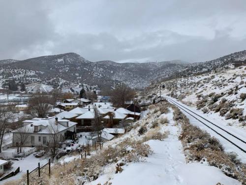 Snow-covered landscape with houses and a railway track, surrounded by mountains under a cloudy sky.
