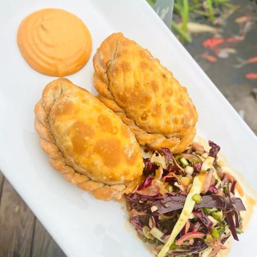 Two golden empanadas on a white plate, accompanied by a colorful slaw and a dollop of orange sauce.