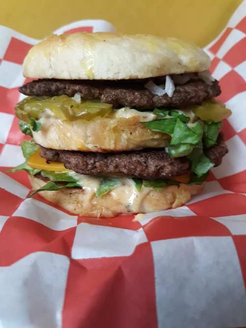 A tall burger stacked with two beef patties, lettuce, pickles, and sauce, served in a checkered paper wrapper.