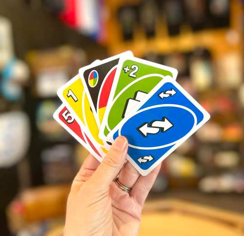 A hand holding five colorful Uno cards, including a +2 and a reverse card, against a blurred background.