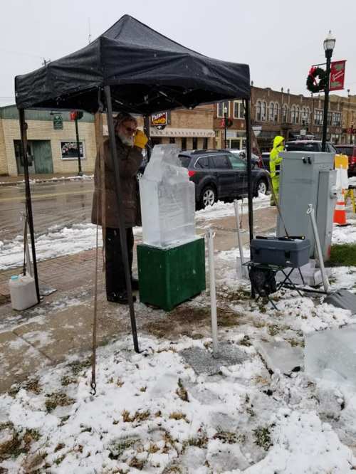 A person carves an ice sculpture under a tent in a snowy outdoor setting, with cars and holiday decorations in the background.