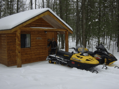 A wooden cabin in a snowy forest, with two parked snowmobiles in front and a person standing by the entrance.
