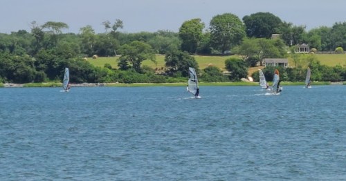 Windsurfers glide across a calm blue lake, surrounded by lush greenery and distant houses on a sunny day.