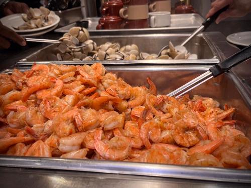 A buffet tray filled with cooked shrimp, with another tray of clams in the background. Tongs are used for serving.