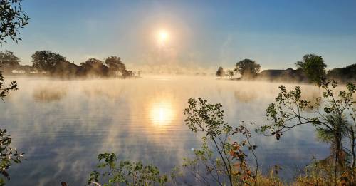 A serene lake at sunrise, with mist rising from the water and trees silhouetted against the bright sky.