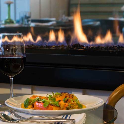 A glass of red wine beside a plate of colorful salad, with a fireplace glowing in the background.