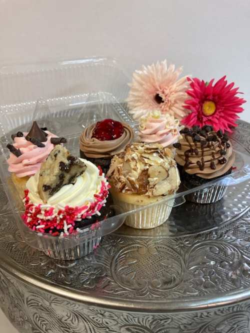 A variety of colorful cupcakes in a clear container, topped with frosting, sprinkles, and flowers.
