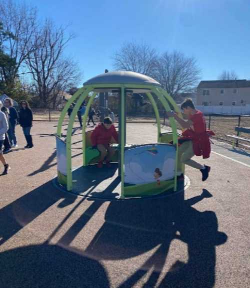 Two children play on a colorful spinning structure at a playground, surrounded by trees and other kids in the background.