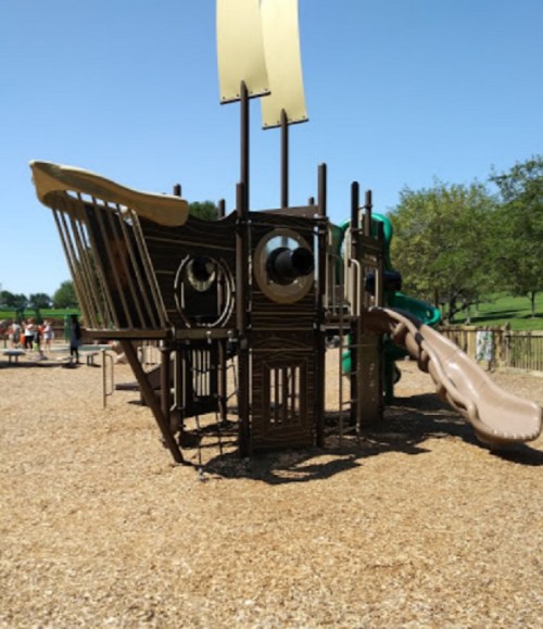 A playground structure resembling a pirate ship, featuring slides and climbing areas, set in a sunny park.