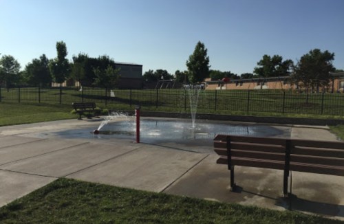 A splash pad with water jets, surrounded by benches and grassy areas, under a clear blue sky.