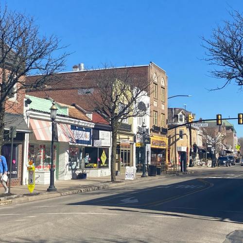 A sunny street scene with shops and trees, featuring clear blue skies and minimal pedestrian activity.