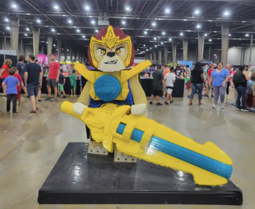 A large LEGO sculpture of a character holding a yellow sword, with a crowd of people in the background at an event.