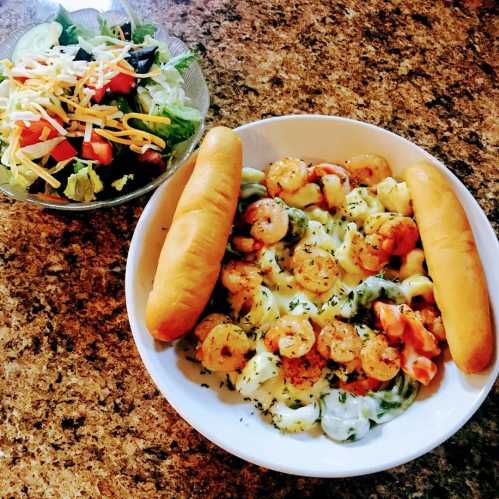 A bowl of shrimp pasta with creamy sauce, garnished with herbs, alongside a fresh salad and two breadsticks.