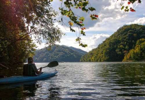 A person kayaking on a calm river surrounded by lush green hills and a partly cloudy sky.