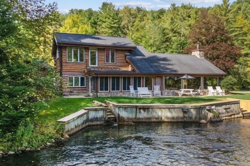 A rustic log cabin by a serene lake, surrounded by lush greenery and featuring a porch with rocking chairs.
