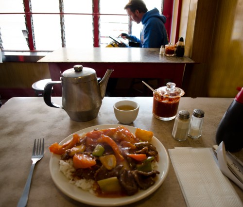 A plate of beef stir-fry with vegetables and rice, a teapot, and condiments on a table, with a person in the background.