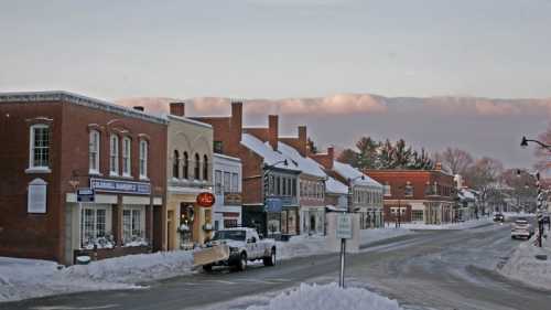 Snow-covered street lined with historic brick buildings and shops, under a cloudy sky. A truck drives down the road.