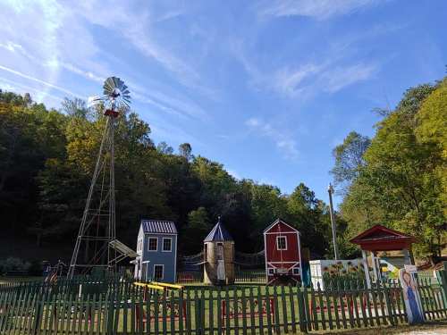 A colorful playground with a windmill, play structures, and a green fence, surrounded by trees and a blue sky.