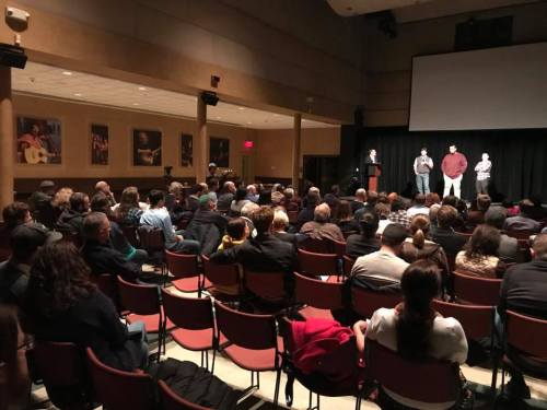 A large audience seated in a theater, watching three speakers on stage during an event.