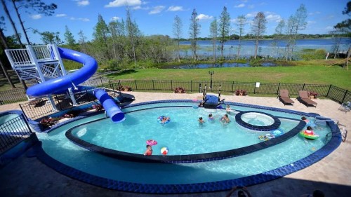 A pool with a blue slide, surrounded by lounge chairs and people enjoying the water, with a scenic lake in the background.