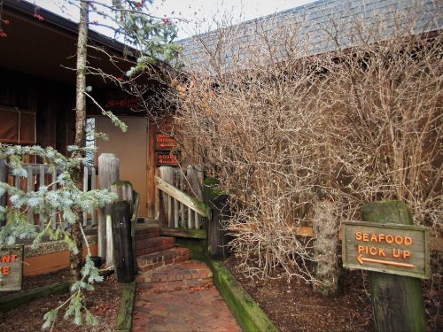 A rustic entrance with wooden steps, surrounded by bare bushes and trees, featuring a sign for seafood pickup.