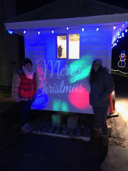 Two women stand outside a brightly lit cabin with a "Merry Christmas" projection and colorful lights.