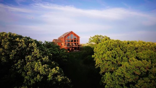 A large wooden house surrounded by lush green trees under a clear blue sky.