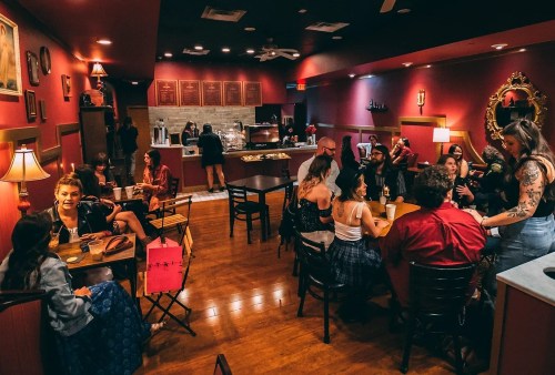 A cozy café scene with people socializing, warm lighting, and a mix of tables and chairs. Snacks are visible in the background.