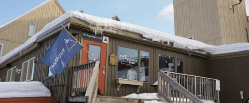 A cozy shop with a wooden exterior, snow on the roof, and a blue flag outside, set against a clear sky.