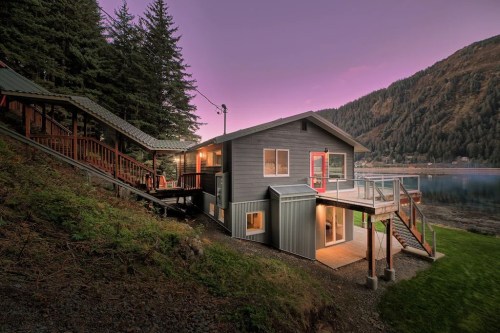 A modern lakeside cabin at dusk, surrounded by trees and mountains, with a red door and warm lights glowing inside.