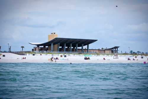 A beach scene featuring a modern building on the shore, with people enjoying the sand and water under a cloudy sky.