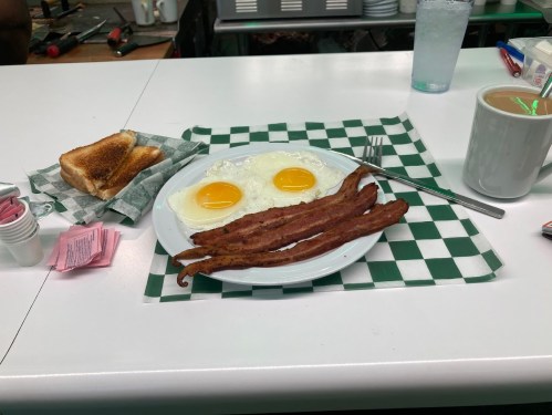 A plate with two sunny-side-up eggs, crispy bacon, toast, and a cup of coffee on a checkered tablecloth.